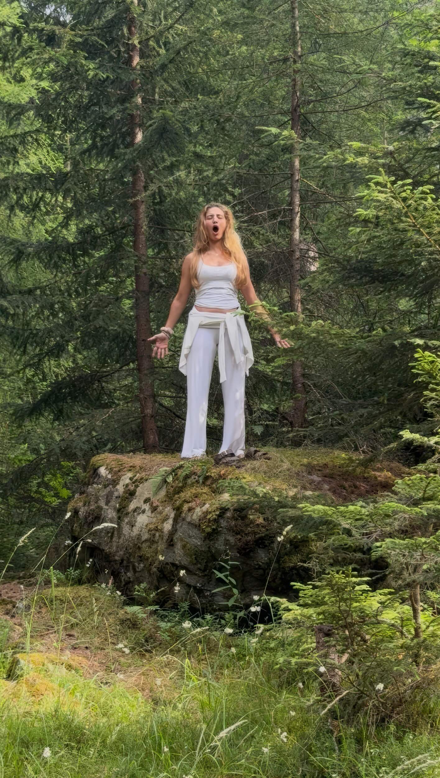 Una donna vestita di bianco canta in piedi su una roccia in mezzo a un bosco di abeti