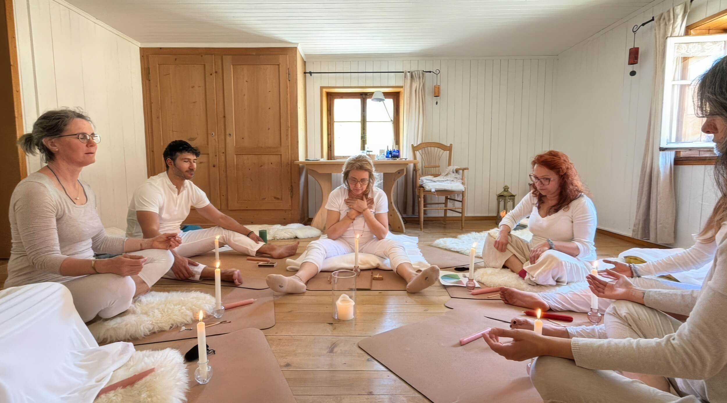 Gruppo vestito di bianco seduto in cerchio su tappetini, con candele accese, durante una meditazione in una stanza rivestita di legno chiaro