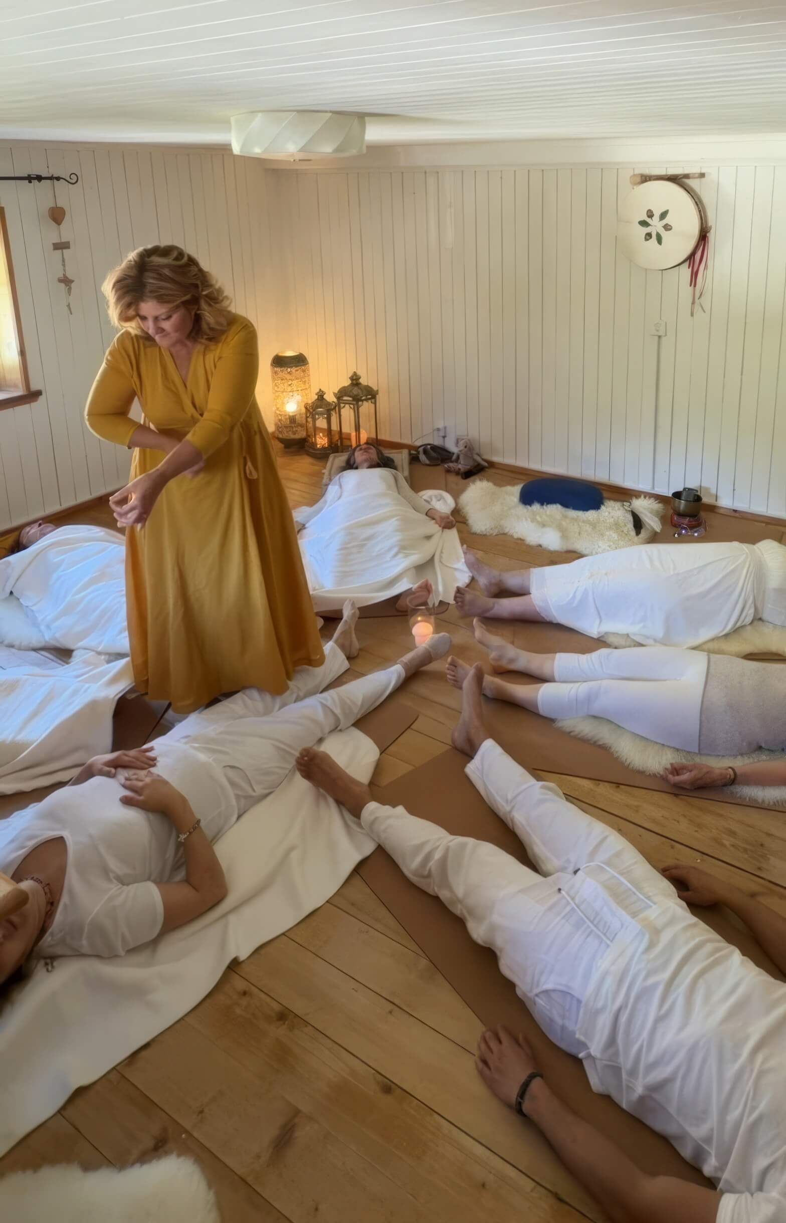 Una facilitatrice in abito ocra guida una pratica mentre i partecipanti, vestiti di bianco, sono sdraiati sul pavimento in legno