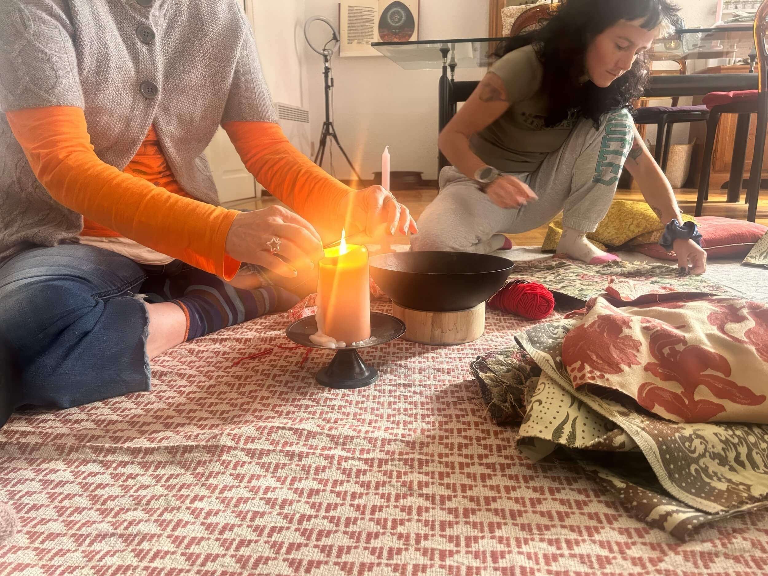 Due donne preparano un rituale sul pavimento con una candela accesa, una ciotola scura e tessuti decorati